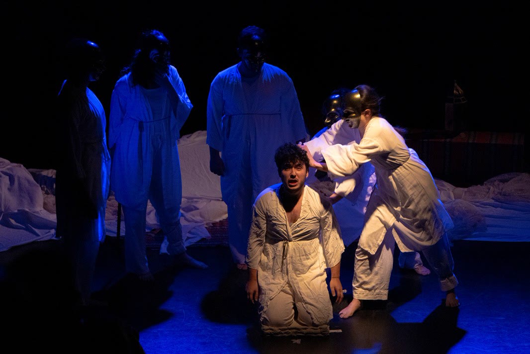 A performer kneels in a pool of blue light while masked figures in white stand around him
