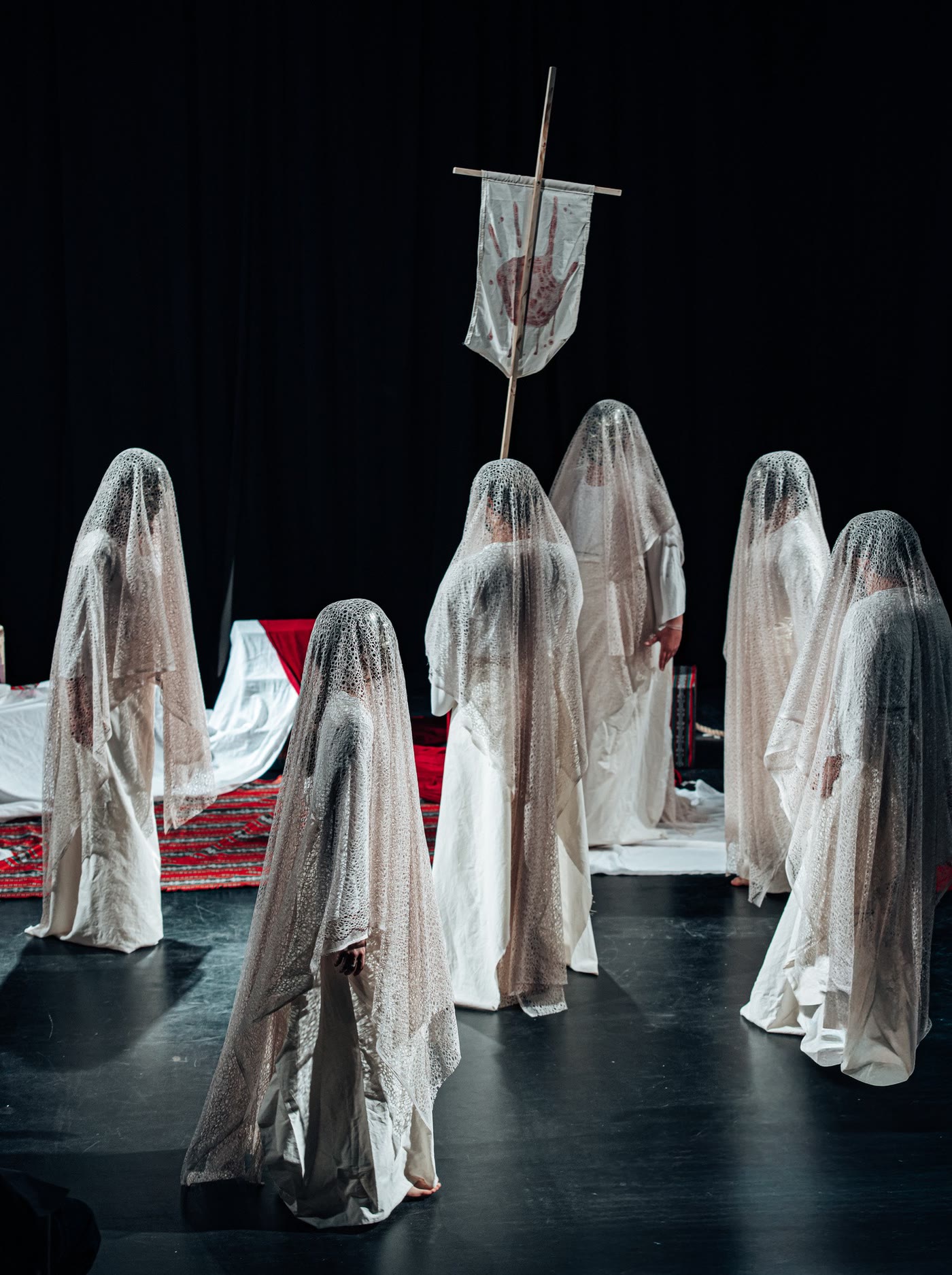 Veiled performers in white lace stand around a banner marked with a red handprint
