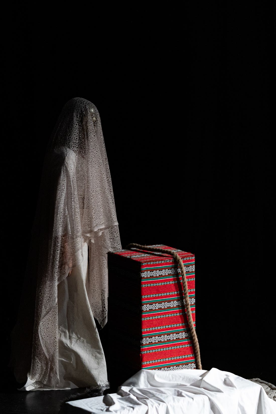 A lone figure draped in white lace stands beside a red woven box on a dark stage
