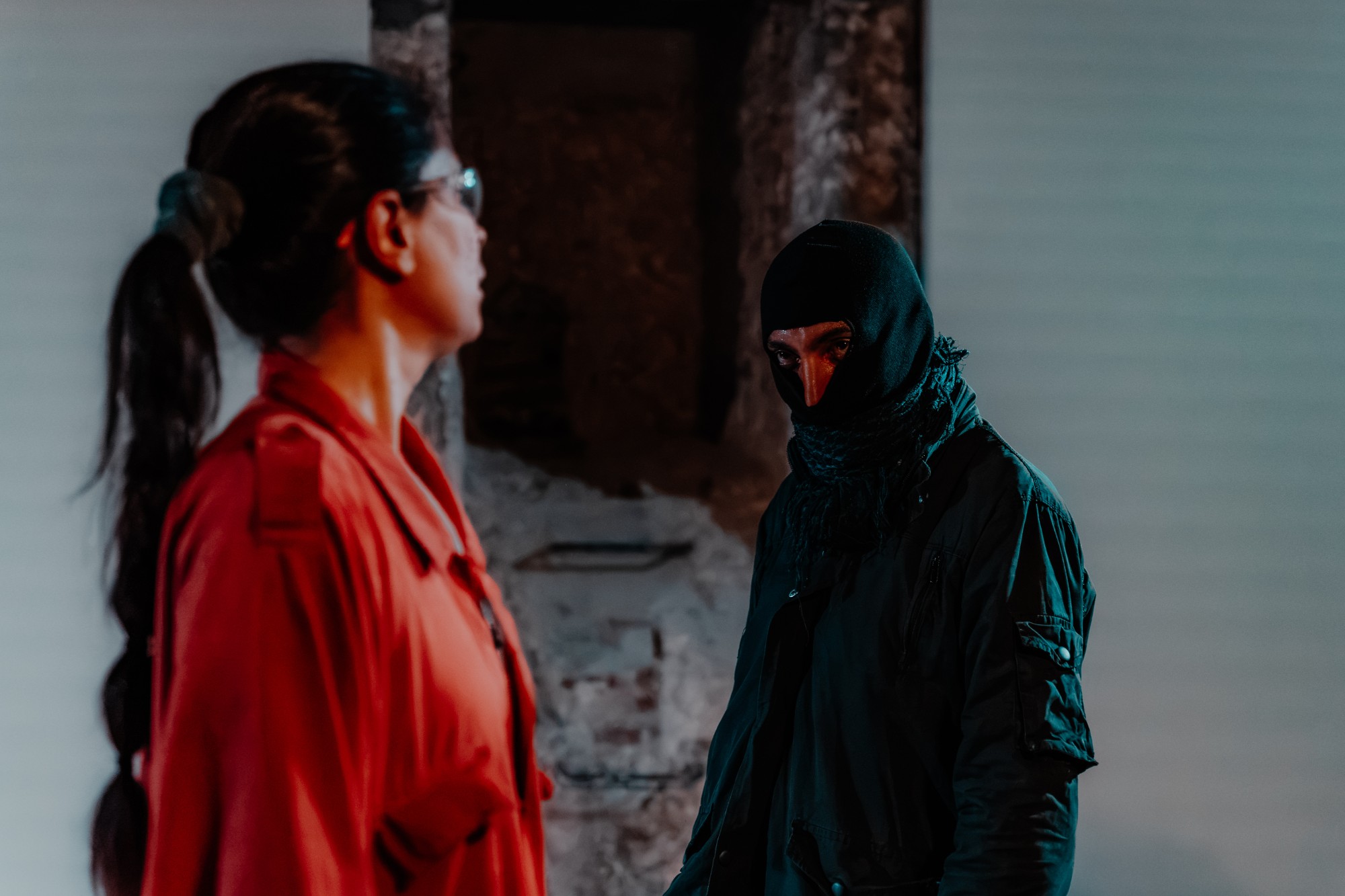 A performer in a balaclava and dark jacket stares past a woman in a red jumpsuit